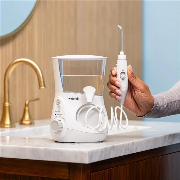 A hand holding the flossing wand of a white Waterpik Aquarius water flosser beside a bathroom sink.