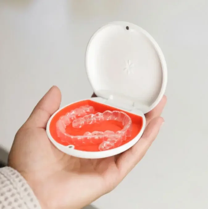 A hand holding an open white Invisalign case with an orange interior and clear aligner trays inside.