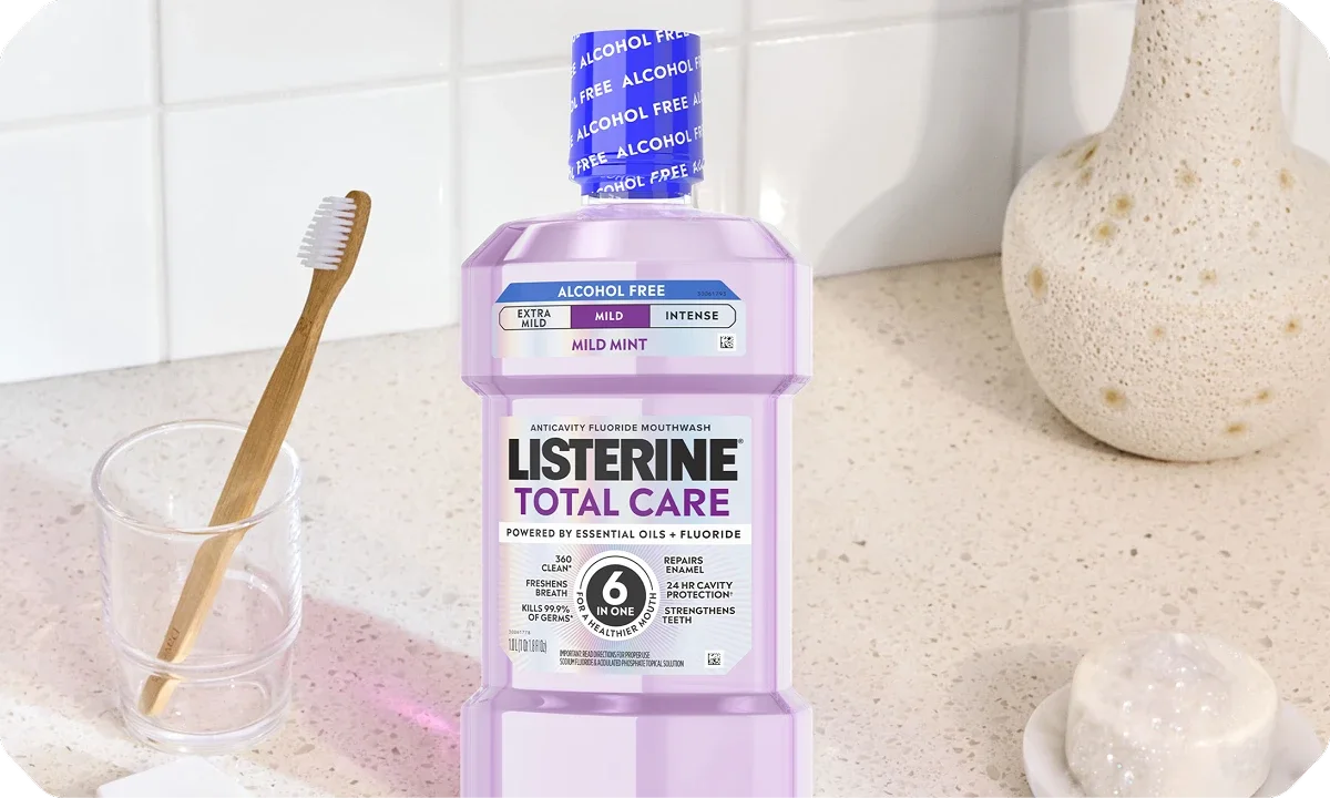 A purple bottle of Listerine Total Care Mild Mint mouthwash beside a wooden toothbrush in a glass on a bathroom counter.
