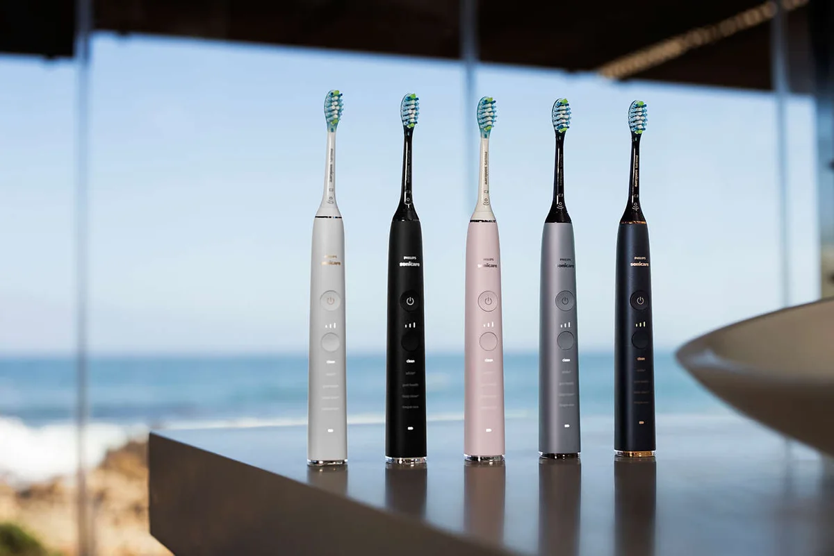 Five Philips Sonicare electric toothbrushes in white, black, pink, grey and dark blue standing on a bathroom counter.